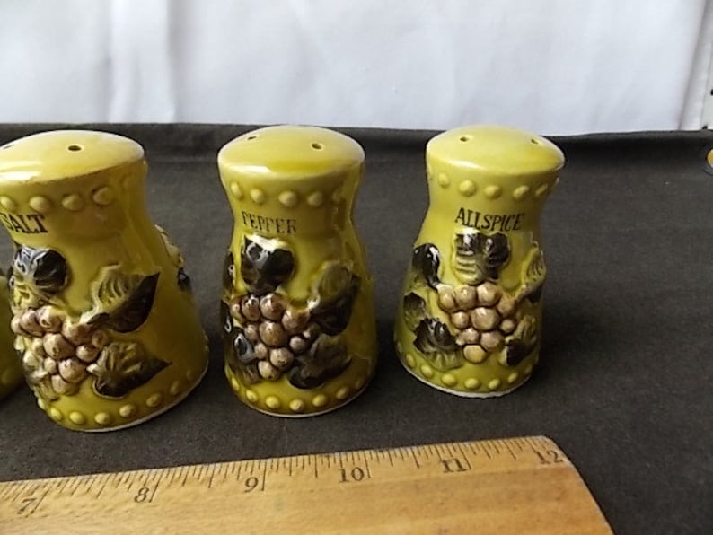May include: A set of three ceramic spice shakers in a light green color. Each shaker is labeled with a spice name: Salt, Pepper, and Allspice. The shakers are decorated with a grape design.