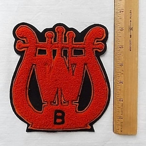 May include: An orange and black patch in the shape of a lyre, with the letter "B" at the bottom. The patch is approximately 5 inches tall. A wooden ruler is next to the patch.