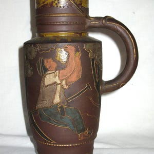 Peut inclure: Une grande cruche en c&eacute;ramique brune pr&eacute;sentant une illustration en relief d'une personne assise buvant dans un r&eacute;cipient. La cruche poss&egrave;de une bande jaune moutarde sur le haut avec des &eacute;toiles et une anse courb&eacute;e sur le c&ocirc;t&eacute; droit du corps.