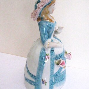 May include: A porcelain figurine of a person wearing a wide-brimmed hat with pink ruffled trim, a blue bodice, and a white bell-shaped skirt decorated with blue vertical stripes, small painted floral accents, and three-dimensional pink roses.