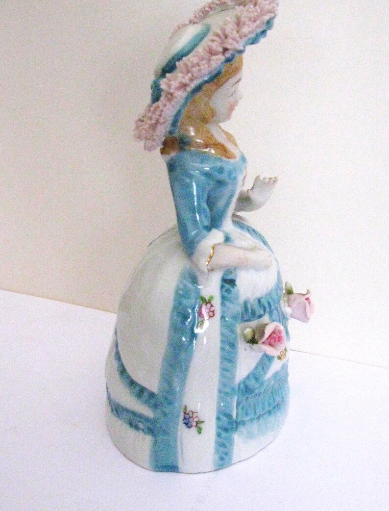 May include: A porcelain figurine of a person wearing a wide-brimmed hat with pink ruffled trim, a blue bodice, and a white bell-shaped skirt decorated with blue vertical stripes, small painted floral accents, and three-dimensional pink roses.