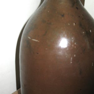 Pode incluir: Um jarro de gr&eacute;s castanho com uma pega. O jarro tem um corpo arredondado e um gargalo estreito. A superf&iacute;cie tem um acabamento brilhante com algumas imperfei&ccedil;&otilde;es. O jarro &eacute; provavelmente uma pe&ccedil;a antiga ou vintage.