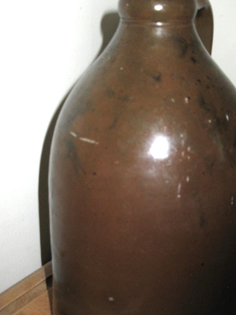 Pode incluir: Um jarro de gr&eacute;s castanho com uma pega. O jarro tem um corpo arredondado e um gargalo estreito. A superf&iacute;cie tem um acabamento brilhante com algumas imperfei&ccedil;&otilde;es. O jarro &eacute; provavelmente uma pe&ccedil;a antiga ou vintage.