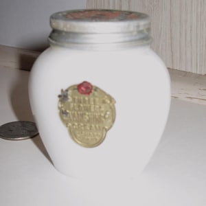 May include: A small, white, vintage jar with a silver lid and a gold label. The label features the text "Three Flowers Vanishing Cream" and a red rose. The jar is likely for cosmetics or skincare products.