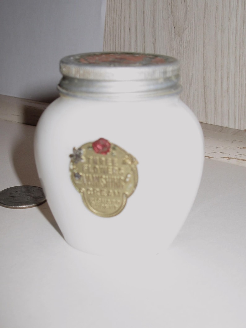 May include: A small, white, vintage jar with a silver lid and a gold label. The label features the text "Three Flowers Vanishing Cream" and a red rose. The jar is likely for cosmetics or skincare products.