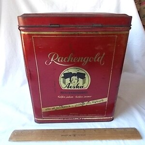 May include: A rectangular red metal tin with a hinged lid, featuring the text "Rachengold" and "Aeska" with a graphic of a person's face. A wooden ruler is placed in front of the tin, showing a length of approximately 12 inches for scale.