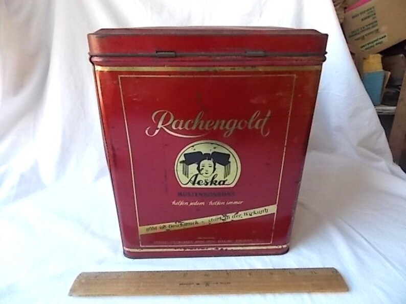 May include: A rectangular red metal tin with a hinged lid, featuring the text "Rachengold" and "Aeska" with a graphic of a person's face. A wooden ruler is placed in front of the tin, showing a length of approximately 12 inches for scale.