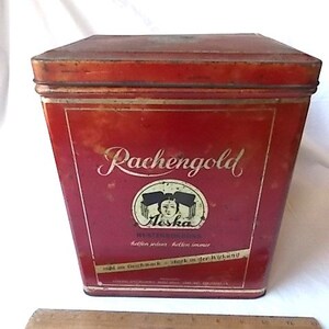 May include: A rectangular red metal tin with a lid, featuring the text Rachengold and Aeska with a graphic of a person. A wooden ruler is placed in front of the tin, showing a measurement of approximately 8 inches in length for the ruler.
