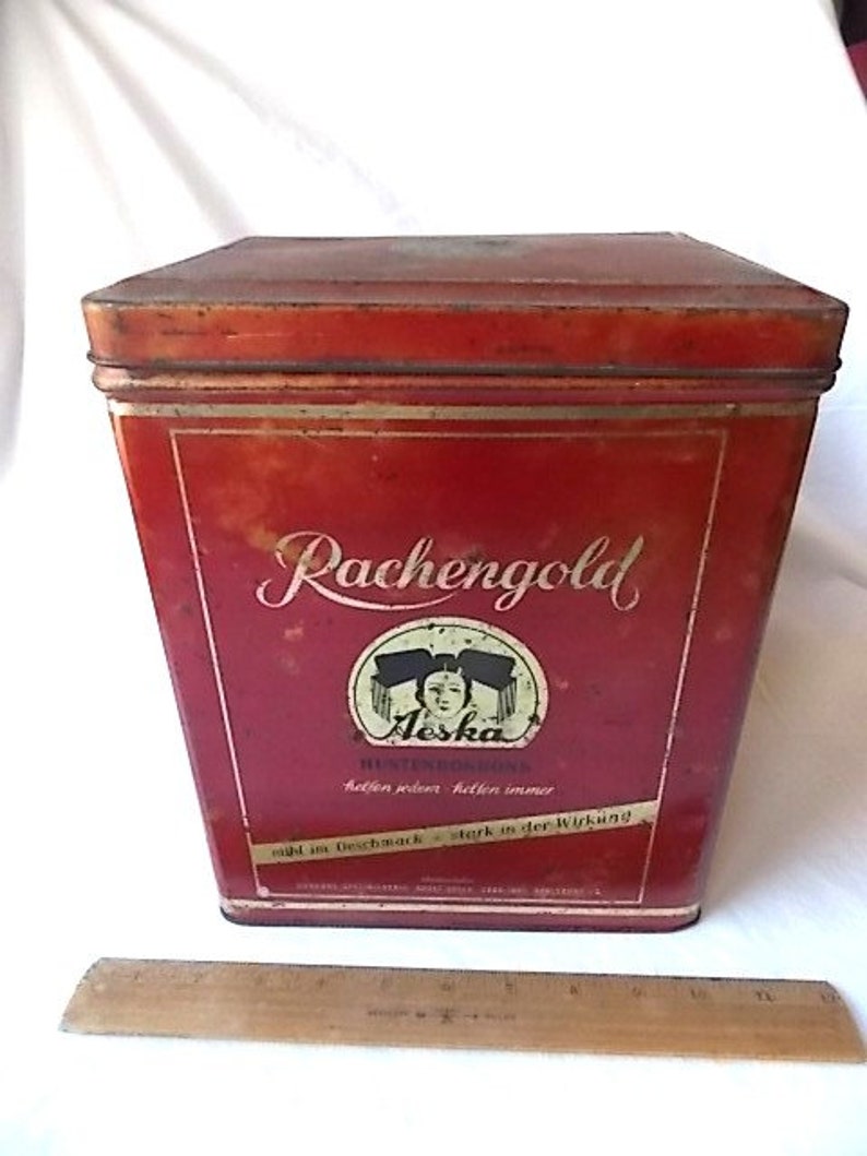 May include: A rectangular red metal tin with a lid, featuring the text Rachengold and Aeska with a graphic of a person. A wooden ruler is placed in front of the tin, showing a measurement of approximately 8 inches in length for the ruler.
