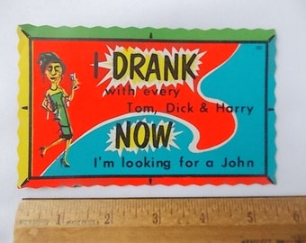 May include: A rectangular vintage postcard with scalloped edges featuring a cartoon illustration of a person in a green dress holding a glass. The text reads: I DRANK with every Tom, Dick & Harry NOW I'm looking for a John. A ruler shows it is 5.5 inches wide.