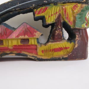 Peut inclure: Une pi&egrave;ce d&eacute;corative en bois sculpt&eacute; &agrave; la main pr&eacute;sentant un paysage d&eacute;coup&eacute;. Le motif comprend des maisons jaunes et rouges aux toits pointus, des palmiers verts et une base brun fonc&eacute;. Le bois pr&eacute;sente des signes d'usure avec de la peinture &eacute;caill&eacute;e.