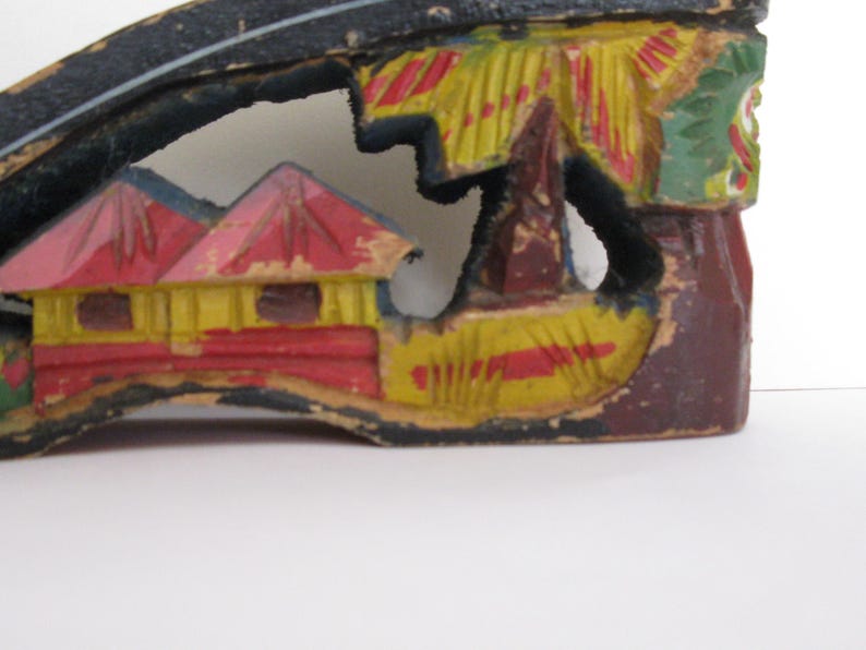 Peut inclure: Une pi&egrave;ce d&eacute;corative en bois sculpt&eacute; &agrave; la main pr&eacute;sentant un paysage d&eacute;coup&eacute;. Le motif comprend des maisons jaunes et rouges aux toits pointus, des palmiers verts et une base brun fonc&eacute;. Le bois pr&eacute;sente des signes d'usure avec de la peinture &eacute;caill&eacute;e.