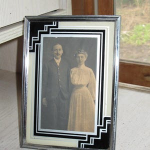 May include: A vintage framed photograph featuring a man and woman in formal attire. The frame has a silver metal border with black and white geometric accents. The photograph is in black and white.