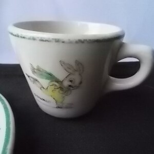 May include: A white ceramic teacup with a small handle and a cartoon rabbit illustration. The cup has a green rim and a matching saucer is partially visible. The rabbit is wearing a yellow outfit and green wings.