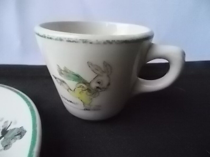May include: A white ceramic teacup with a small handle and a cartoon rabbit illustration. The cup has a green rim and a matching saucer is partially visible. The rabbit is wearing a yellow outfit and green wings.