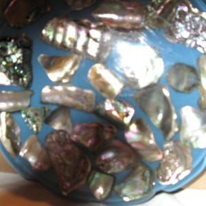 May include: A blue, scalloped-edge decorative bowl decorated with iridescent abalone shell pieces. The shells vary in shape and size, showing silver, purple, and green colours. The bowl's surface is smooth, contrasting with the textured shells.