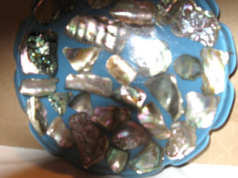 May include: A blue, scalloped-edge decorative bowl decorated with iridescent abalone shell pieces. The shells vary in shape and size, showing silver, purple, and green colours. The bowl's surface is smooth, contrasting with the textured shells.