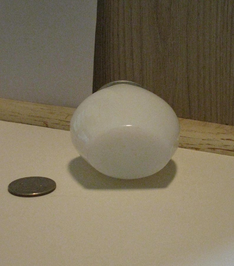 May include: A white, round, glass light fixture with a silver metal ring around the top. A coin is next to the light fixture. The light fixture is on a white surface.