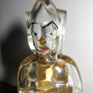 K&ouml;nnte beinhalten: Eine klare Glasfigur einer Figur mit spitzem Kopf, schwarzen Augenbrauen und rotem Mund. Die Figur hat einen goldfarbenen Torso und ist wahrscheinlich ein Dekorations- oder Sammlerobjekt.