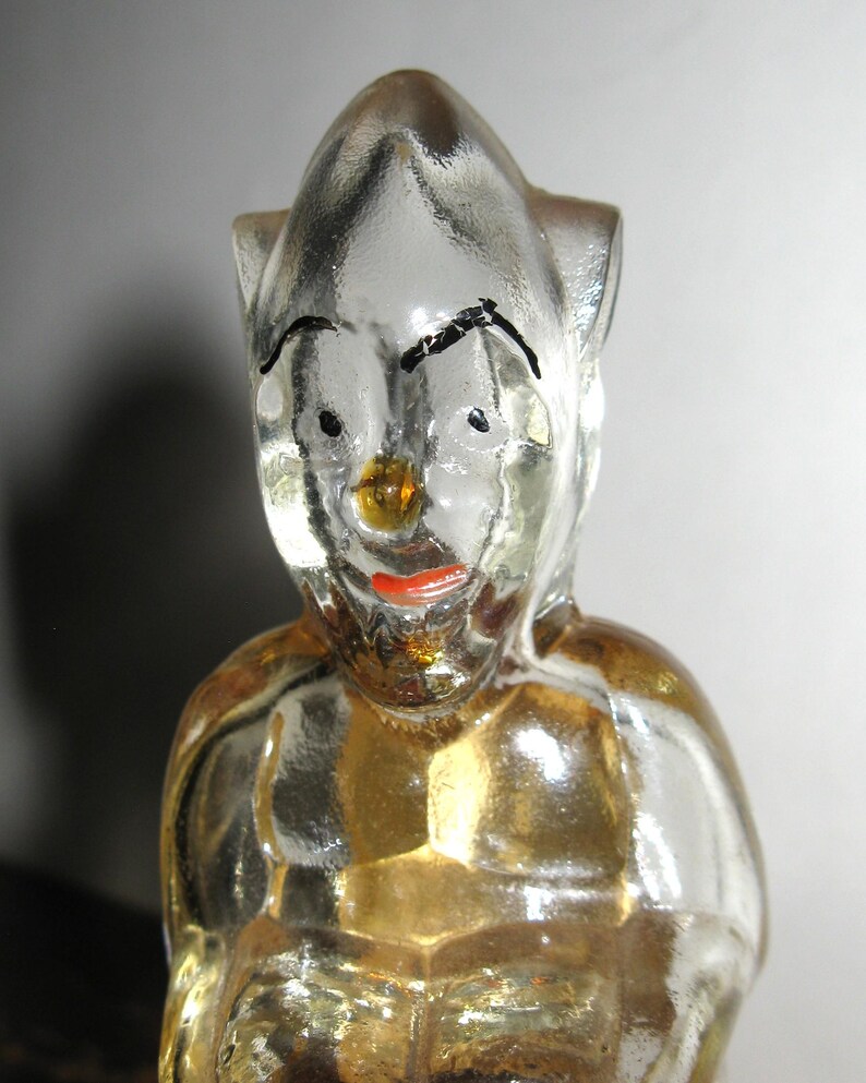 K&ouml;nnte beinhalten: Eine klare Glasfigur einer Figur mit spitzem Kopf, schwarzen Augenbrauen und rotem Mund. Die Figur hat einen goldfarbenen Torso und ist wahrscheinlich ein Dekorations- oder Sammlerobjekt.