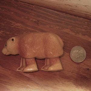 Peut inclure: Un jouet en plastique en forme d'ours brun clair avec quatre pattes, une petite t&ecirc;te et un corps arrondi. Une pi&egrave;ce de monnaie est &agrave; c&ocirc;t&eacute; de l'ours pour la comparaison de taille. L'ours est sur une surface en bois.