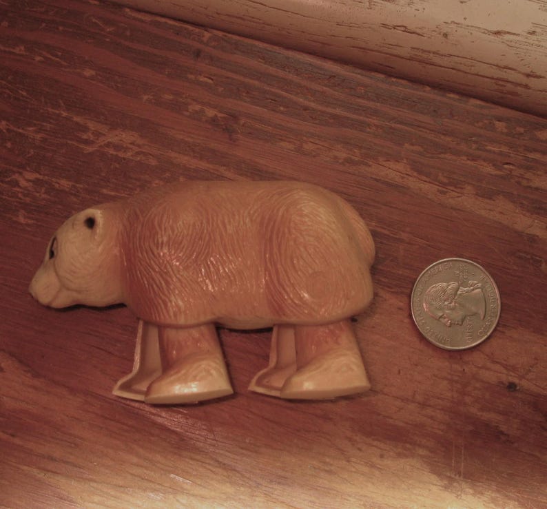 Peut inclure: Un jouet en plastique en forme d'ours brun clair avec quatre pattes, une petite t&ecirc;te et un corps arrondi. Une pi&egrave;ce de monnaie est &agrave; c&ocirc;t&eacute; de l'ours pour la comparaison de taille. L'ours est sur une surface en bois.