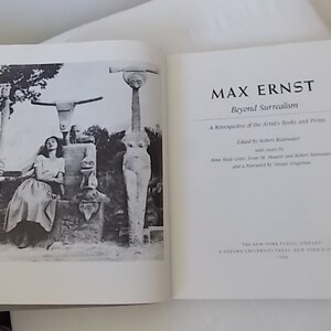 Pode incluir: Livro aberto com o t&iacute;tulo "MAX ERNST Beyond Surrealism" na p&aacute;gina direita. A p&aacute;gina esquerda apresenta uma fotografia a preto e branco de uma mulher e esculturas. Uma retrospectiva dos livros e gravuras do artista.