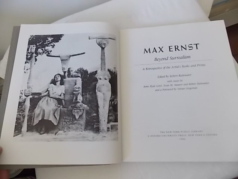 Pode incluir: Livro aberto com o t&iacute;tulo "MAX ERNST Beyond Surrealism" na p&aacute;gina direita. A p&aacute;gina esquerda apresenta uma fotografia a preto e branco de uma mulher e esculturas. Uma retrospectiva dos livros e gravuras do artista.