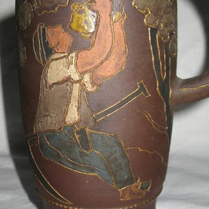 Peut inclure: Une cruche en c&eacute;ramique brune pr&eacute;sentant une illustration en relief d'un personnage portant un chapeau, un gilet et un pantalon, buvant dans un r&eacute;cipient. Le motif inclut des contours dor&eacute;s sur un fond brun mat avec une anse sur le c&ocirc;t&eacute;.