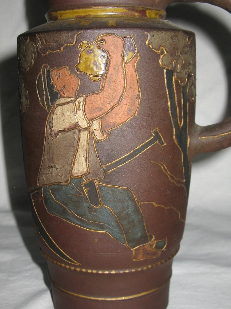 Peut inclure: Une cruche en c&eacute;ramique brune pr&eacute;sentant une illustration en relief d'un personnage portant un chapeau, un gilet et un pantalon, buvant dans un r&eacute;cipient. Le motif inclut des contours dor&eacute;s sur un fond brun mat avec une anse sur le c&ocirc;t&eacute;.