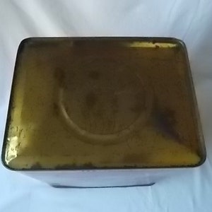 May include: A rectangular metal tin with a weathered, golden-brown finish on its top surface. The lid features a central circular indentation and shows signs of oxidation and wear along the edges, presenting a vintage, aged appearance.