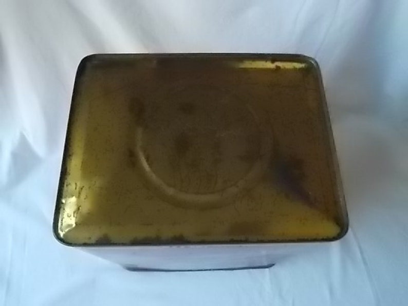 May include: A rectangular metal tin with a weathered, golden-brown finish on its top surface. The lid features a central circular indentation and shows signs of oxidation and wear along the edges, presenting a vintage, aged appearance.