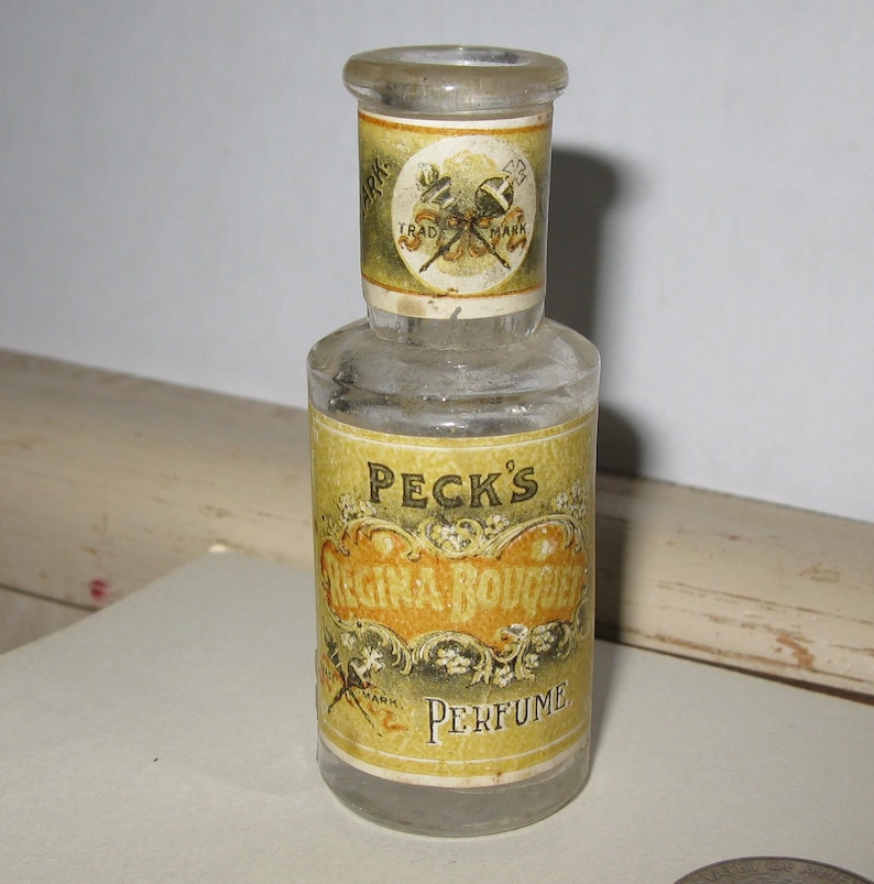Może przedstawiać: Zabytkowa butelka perfum wykonana z przezroczystego szkła, o cylindrycznym kształcie. Butelka posiada ż&oacute;łtą etykietę ze złotymi detalami, z napisem "PECK'S Regina Bouquet Perfume". Etykieta zawiera r&oacute;wnież okrągły emblemat.