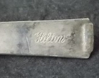 May include: A silver-toned metal spoon handle featuring a smooth, flat surface with the word Hilton embossed in a cursive script font. The metal shows signs of wear and a matte finish, with a slightly raised, rounded end at the top of the handle.