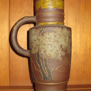 Peut inclure: Une cruche en c&eacute;ramique brune avec une bande textur&eacute;e jaune moutarde sur le haut, orn&eacute;e de petits motifs d'&eacute;toiles et de points. Le corps pr&eacute;sente un relief d'arbres stylis&eacute;s vert p&acirc;le aux contours sombres et des d&eacute;tails dor&eacute;s sur l'anse.