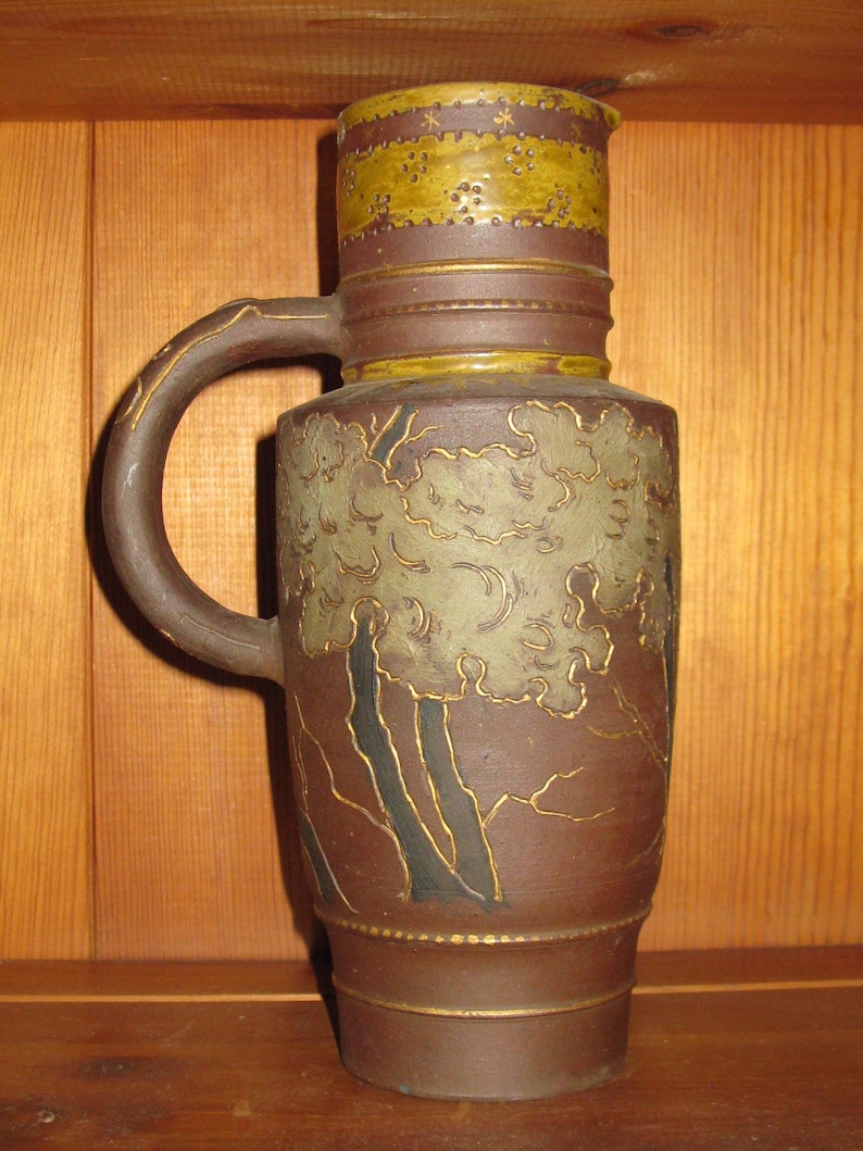 Peut inclure: Une cruche en c&eacute;ramique brune avec une bande textur&eacute;e jaune moutarde sur le haut, orn&eacute;e de petits motifs d'&eacute;toiles et de points. Le corps pr&eacute;sente un relief d'arbres stylis&eacute;s vert p&acirc;le aux contours sombres et des d&eacute;tails dor&eacute;s sur l'anse.
