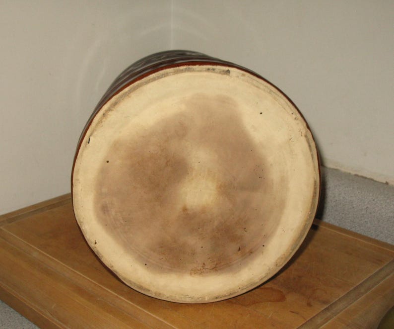 May include: A brown ceramic jug with a wide, circular base and a cylindrical body. The jug has a light brown interior and a darker brown exterior with horizontal ridges. The jug is sitting on a wooden surface.