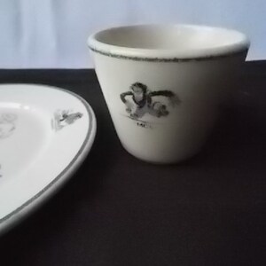 May include: A white ceramic egg cup and a small plate with a gray border. The egg cup has a black and white illustration and the word "MC" on it. The plate has a bunny illustration.