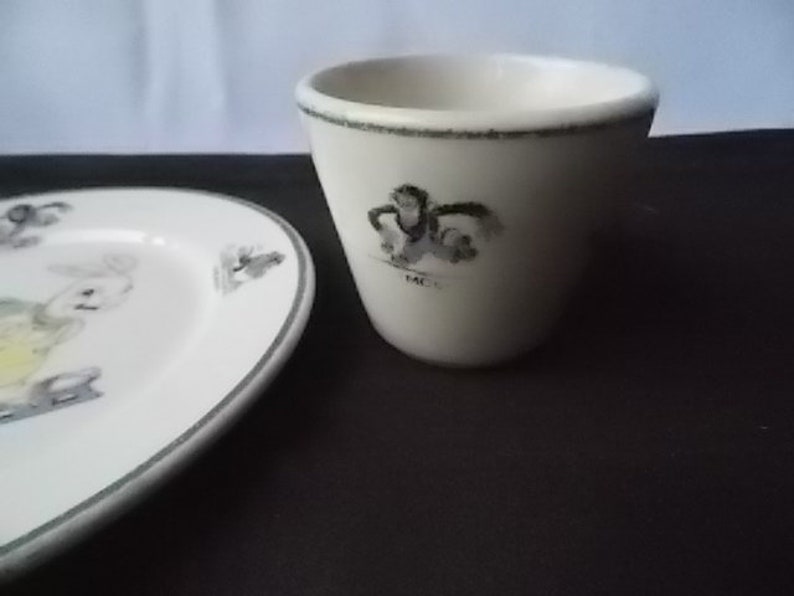 May include: A white ceramic egg cup and a small plate with a gray border. The egg cup has a black and white illustration and the word "MC" on it. The plate has a bunny illustration.