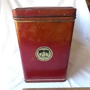 May include: A tall, rectangular red metal tin with a lid, featuring a circular label on the front with the text Aeska Bonbons and an illustration of a person. A wooden ruler is placed in front of the tin to show its scale in inches.