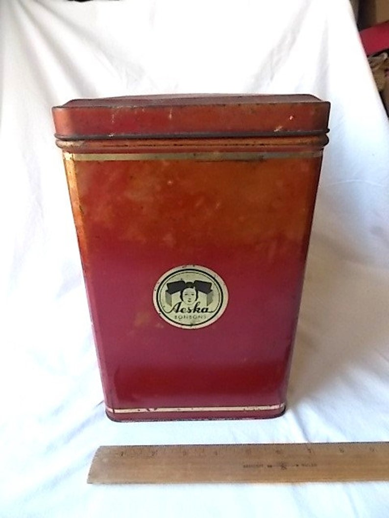 May include: A tall, rectangular red metal tin with a lid, featuring a circular label on the front with the text Aeska Bonbons and an illustration of a person. A wooden ruler is placed in front of the tin to show its scale in inches.