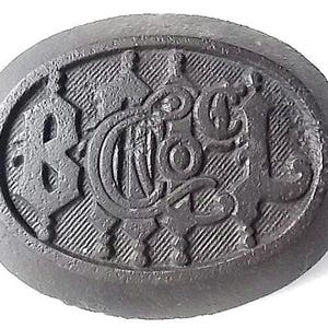 May include: A dark grey, oval button with embossed lettering and decorative details. The button displays the letters "B", "N", "C", and "L" in a stylized font, set against a textured background.