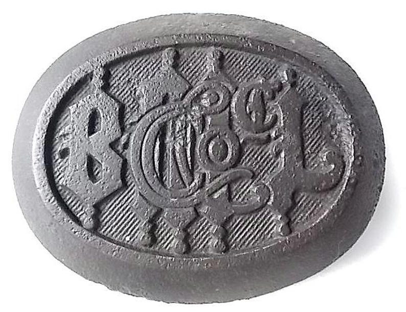 May include: A dark grey, oval button with embossed lettering and decorative details. The button displays the letters "B", "N", "C", and "L" in a stylized font, set against a textured background.