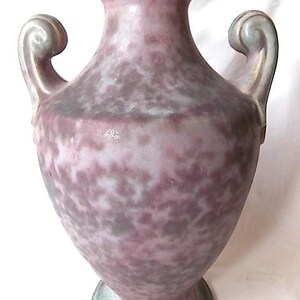 May include: A speckled, antique-style vase in shades of purple, gray, and green. The vase features two curved handles and a flared base. The rim and handles have a gold-colored trim. The vase has a bulbous body and a narrow neck.