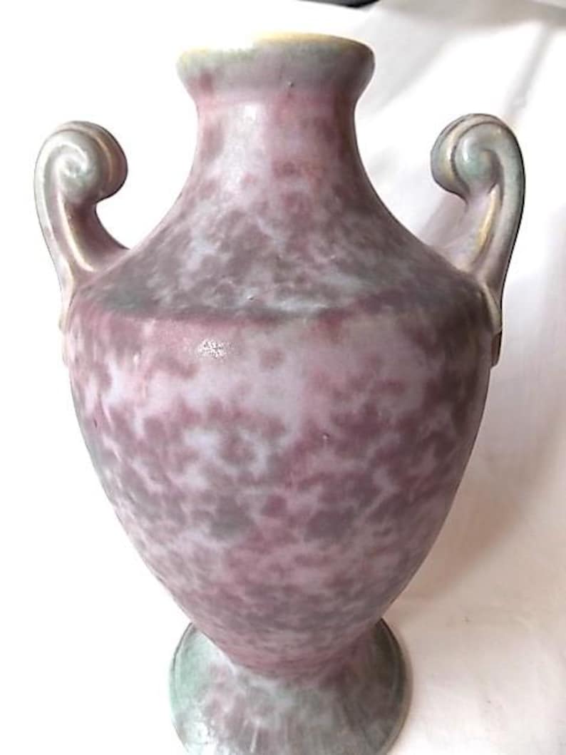 May include: A speckled, antique-style vase in shades of purple, gray, and green. The vase features two curved handles and a flared base. The rim and handles have a gold-colored trim. The vase has a bulbous body and a narrow neck.