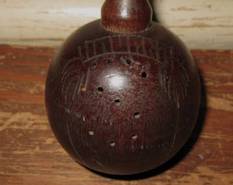 May include: A dark brown, spherical wooden shaker featuring a narrow neck at the top and a series of small, circular holes drilled into the front surface. The wood grain is visible across the rounded body, which also displays faint, etched line details.