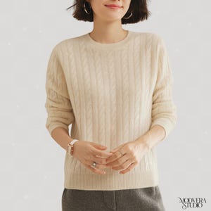Könnte beinhalten: Cremefarbener Zopfstrickpullover mit Rundhalsausschnitt. Der Pullover hat ein strukturiertes Muster und lange Ärmel. Das Model trägt eine weiße Uhr und Ringe. Der Pullover ist neutral und kann mit vielen Outfits kombiniert werden.