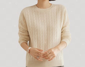 May include: Cream-colored cable knit sweater with a crew neck. The sweater has a textured pattern and long sleeves. The model is wearing a white watch and rings. The sweater is a neutral color and can be paired with many outfits.