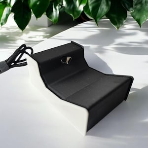 May include: A black charging stand with a white base and a cord. The stand has a small opening on the top for a device to be placed in.