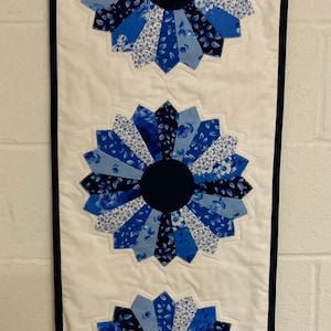May include: A white fabric wall hanging with a navy blue border features three blue and white floral designs. Each design is a circle with a navy blue center and blue and white petals.